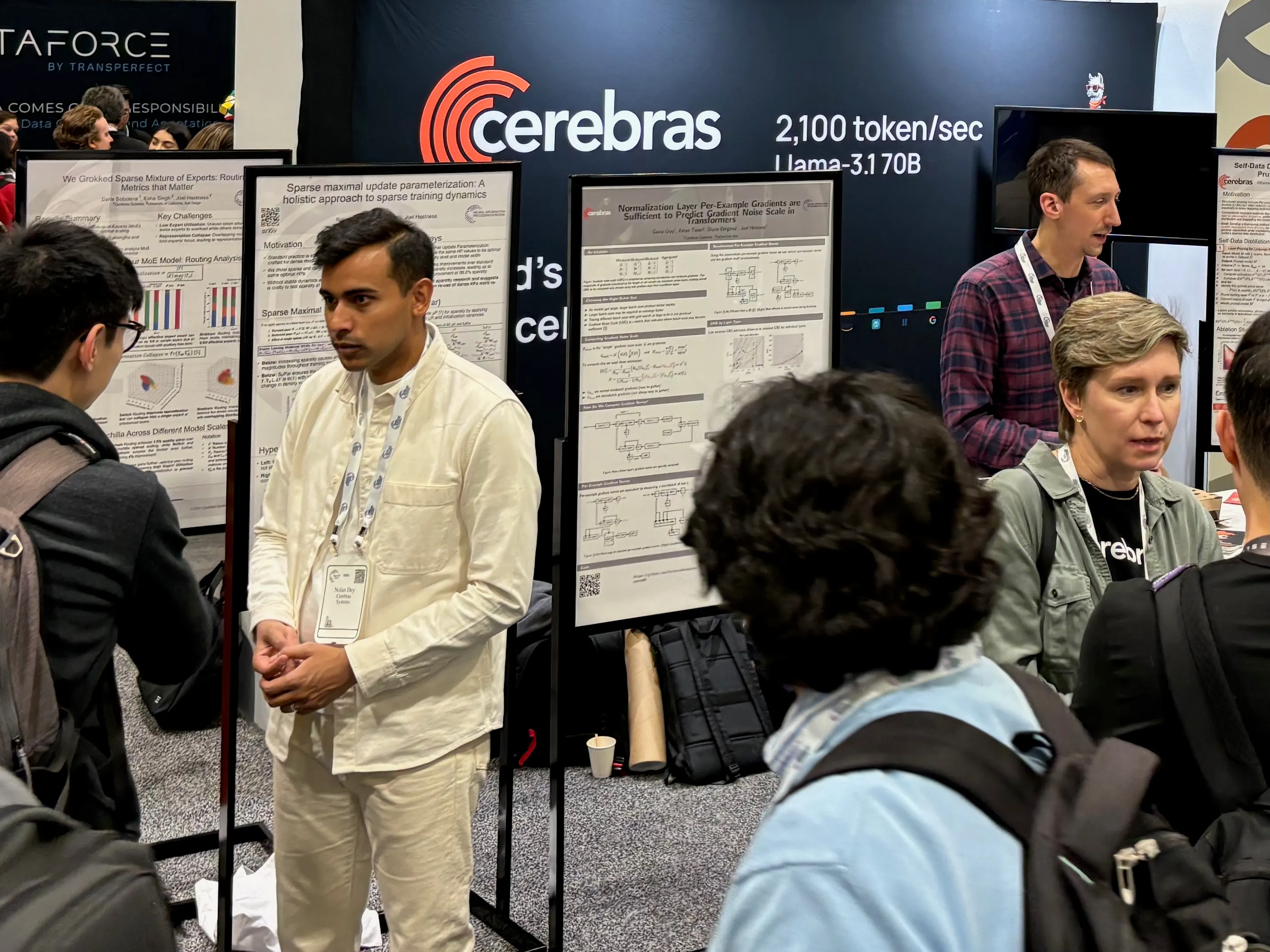 Photo of a sponsor booth at NeurIPS. A banner in the background reads 'cerebras: 2,100 token/sec Llama-3.1 70B'.
Several people are having conversations in the foreground, surrounded by posters containing technical information.