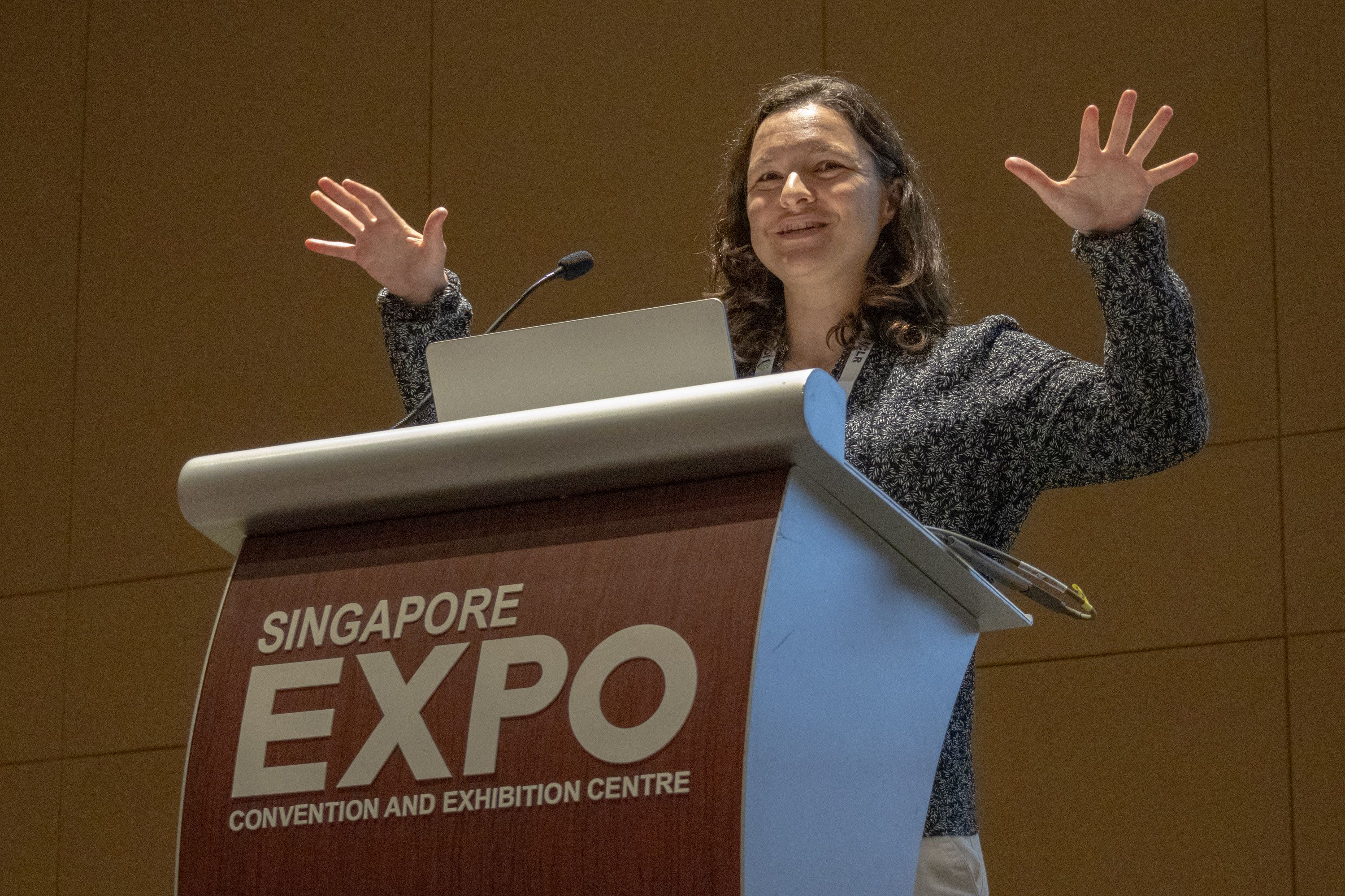 Chelsea Finn addressing the audience from behind a lectern, smiling and holding her hands up in the air. 