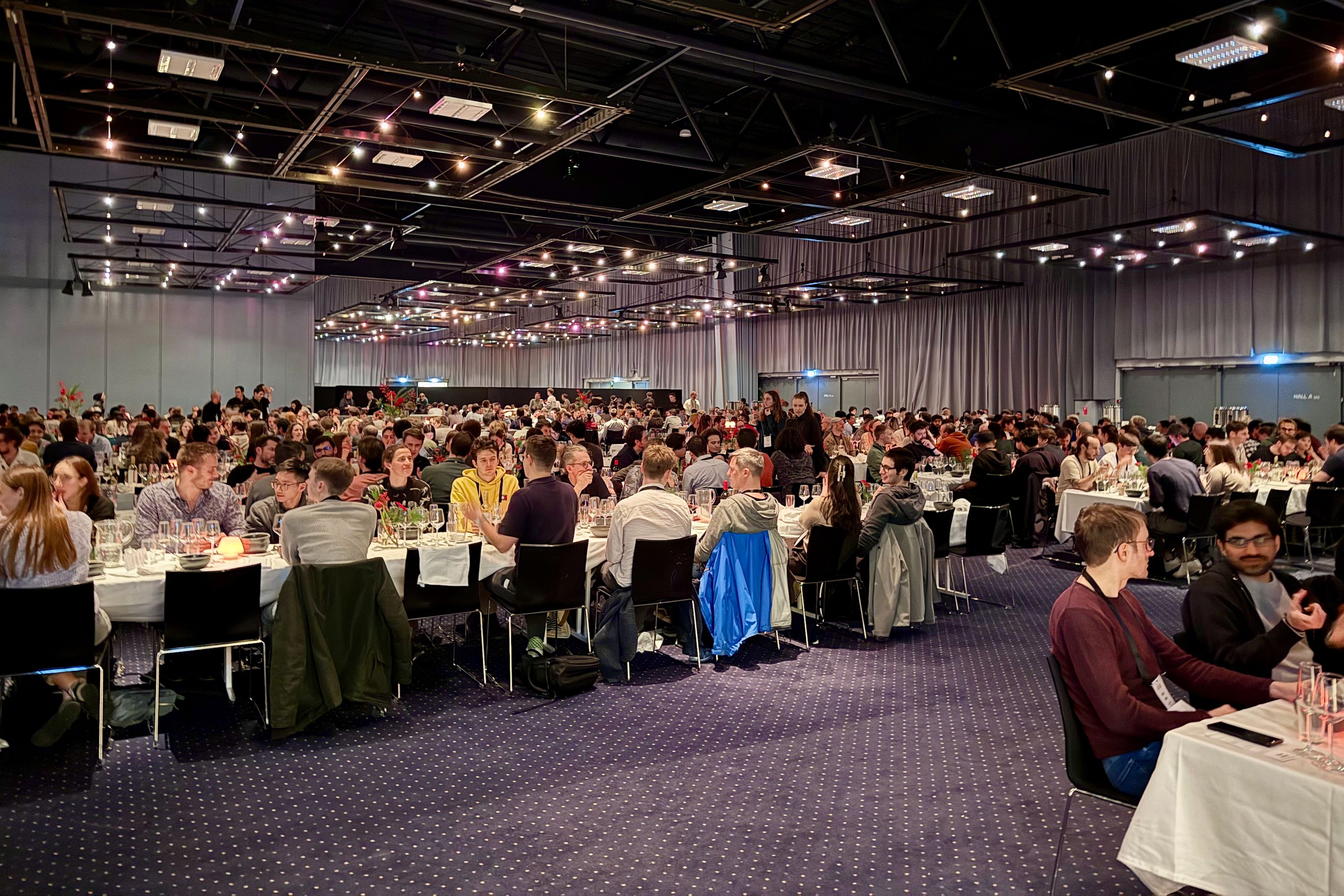 Hundreds of people are seated at long tables in a large hall with overhead lights