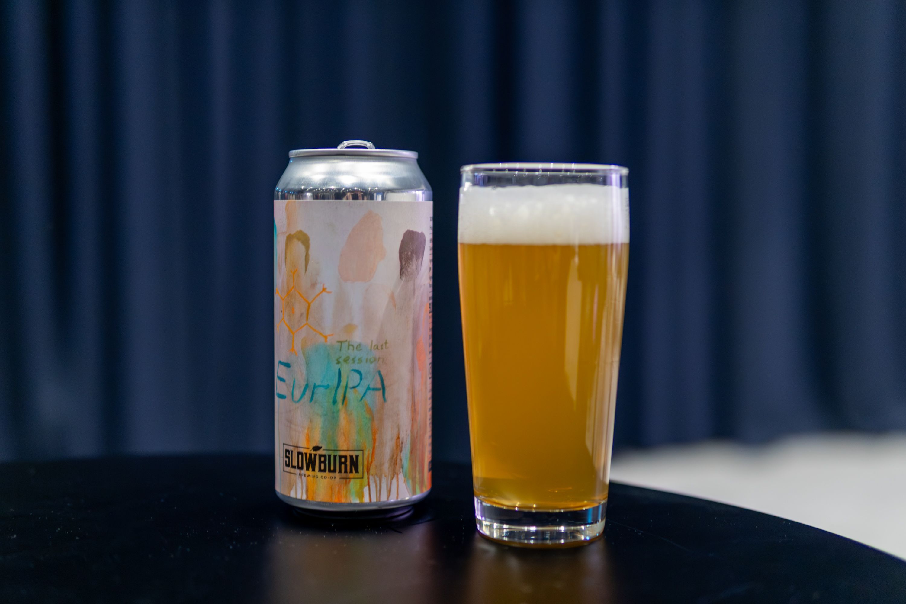 A can of "EurIPA" beer, alongside a glass full of the beer, standing on a small table.