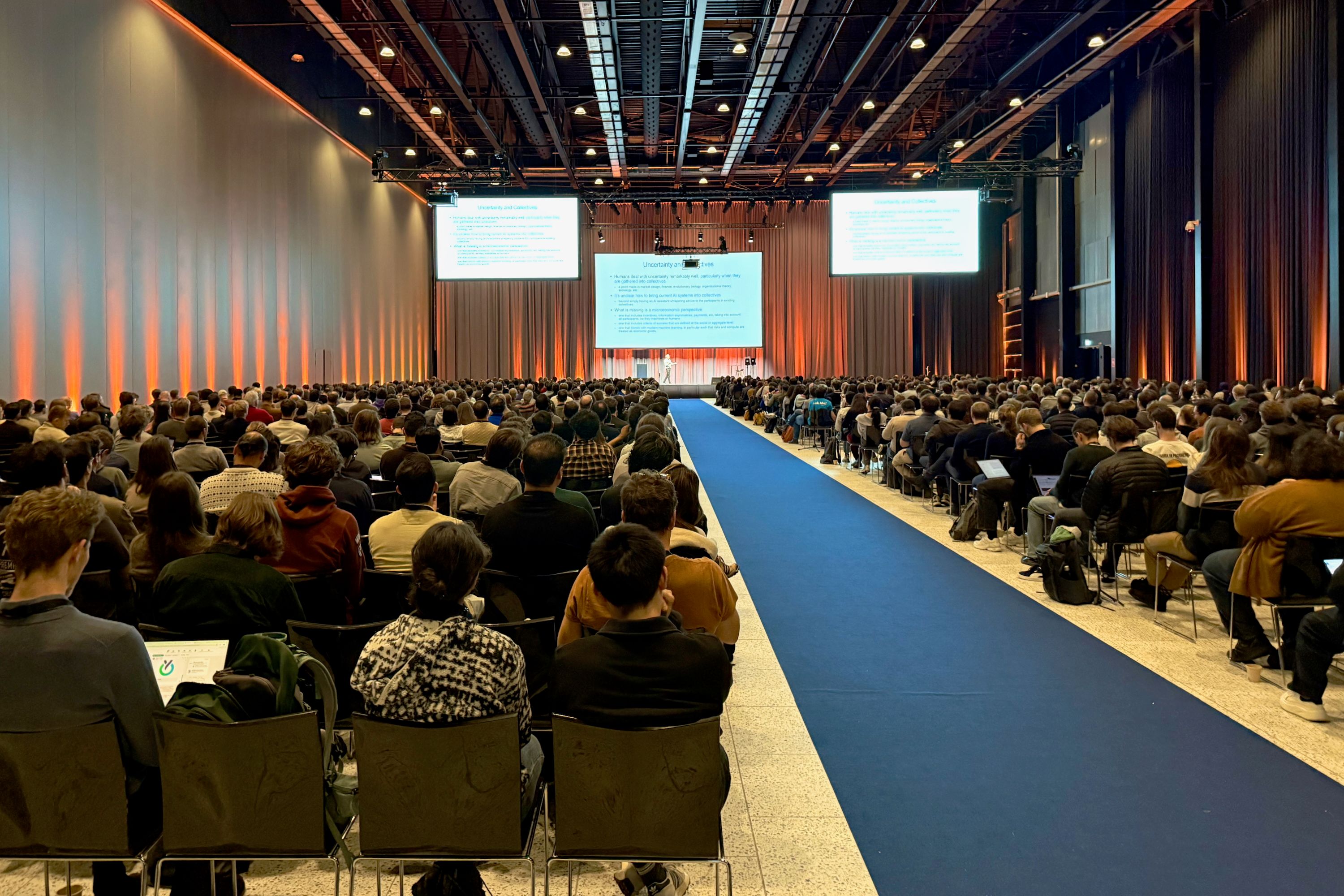 A large hall, with hundreds of people sat down in rows facing a stage with multiple screens. 