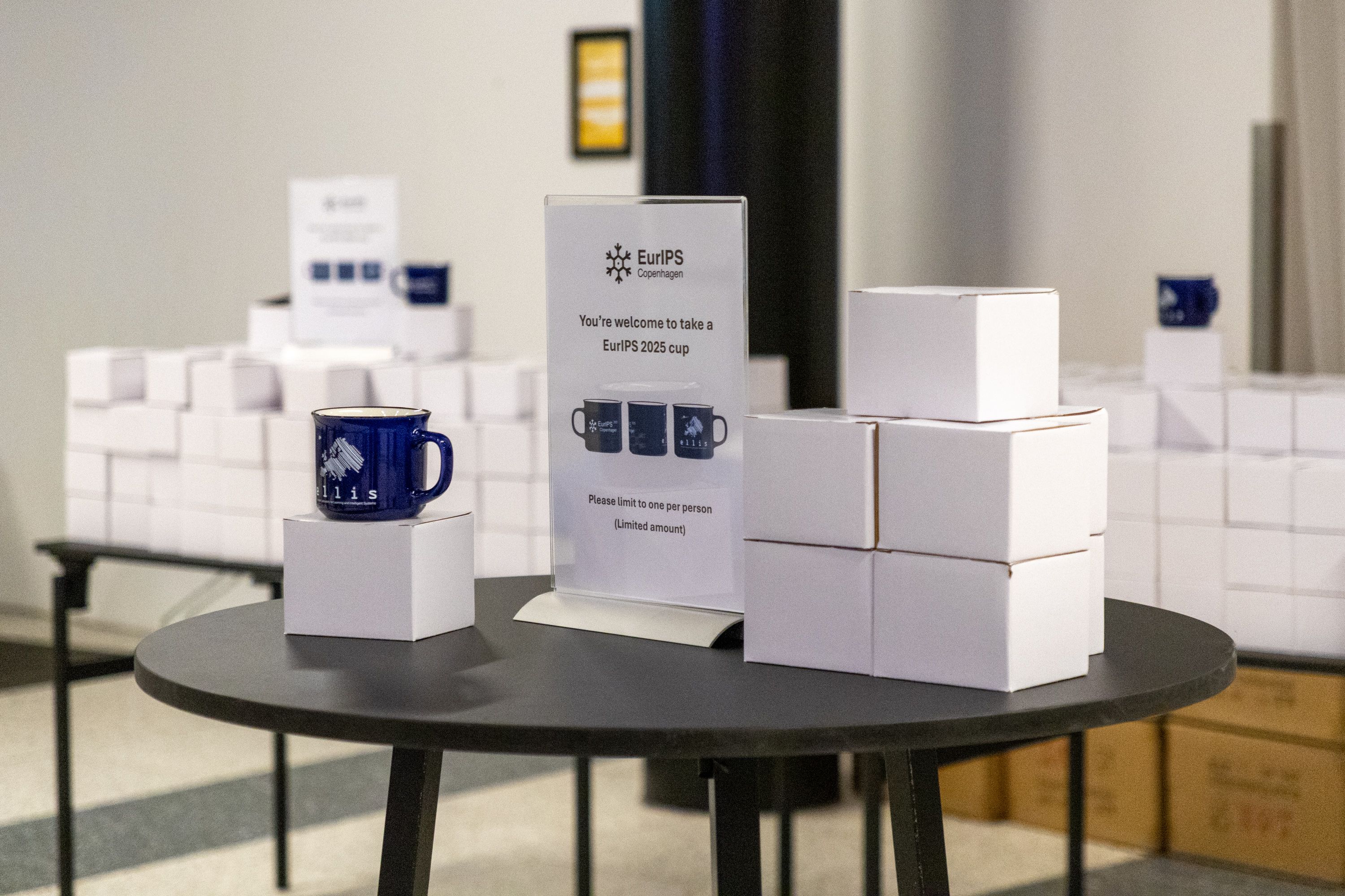 Boxes containing mugs, with a few mugs atop them on display. A sign reads "You're welcome to take a EurIPS 2025 cup". 
