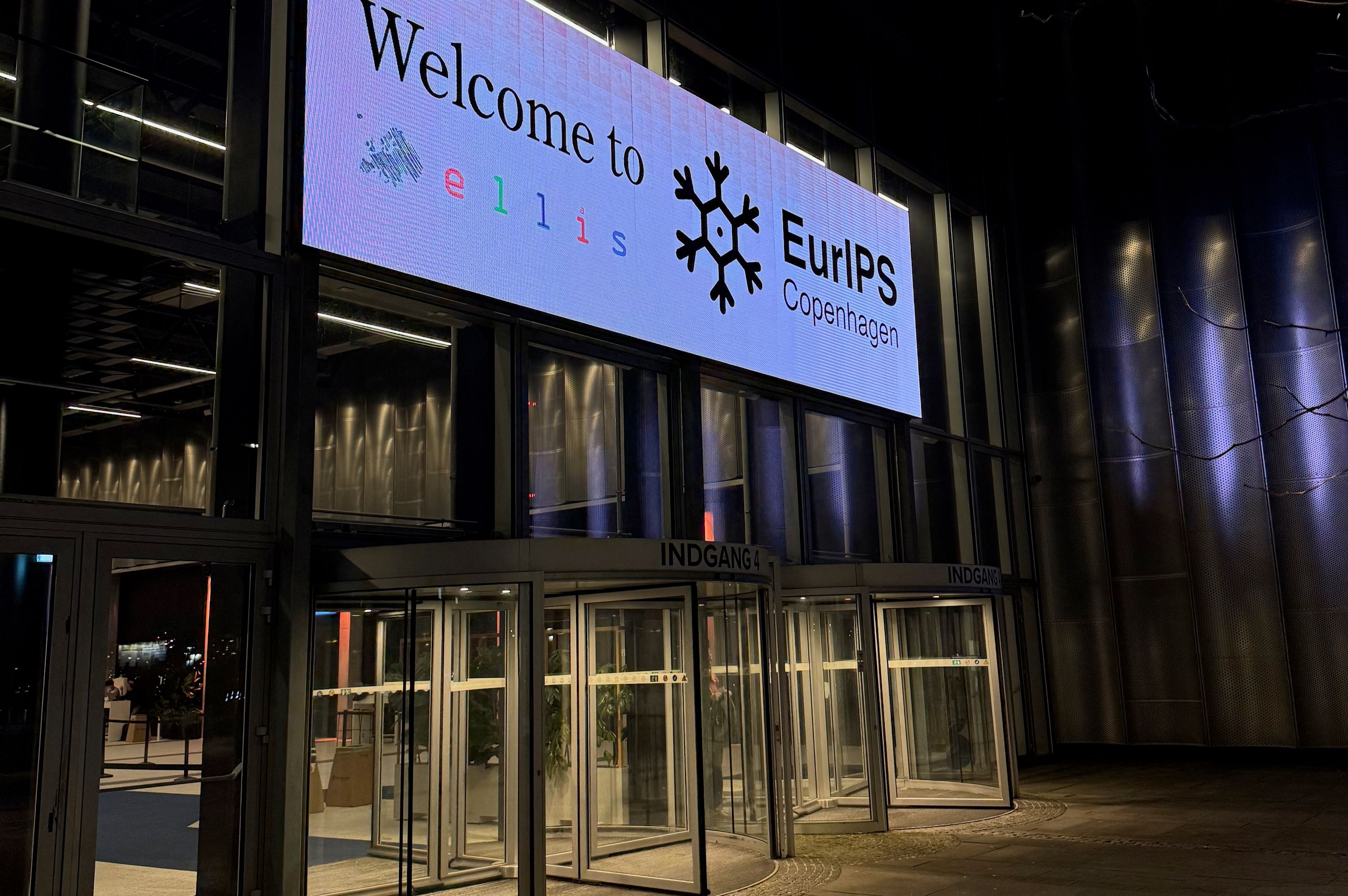 Two revolving doors at the venue entrance, below a sign that reads "Welcome to EurIPS Copenhagen," and bears the ELLIS logo. 