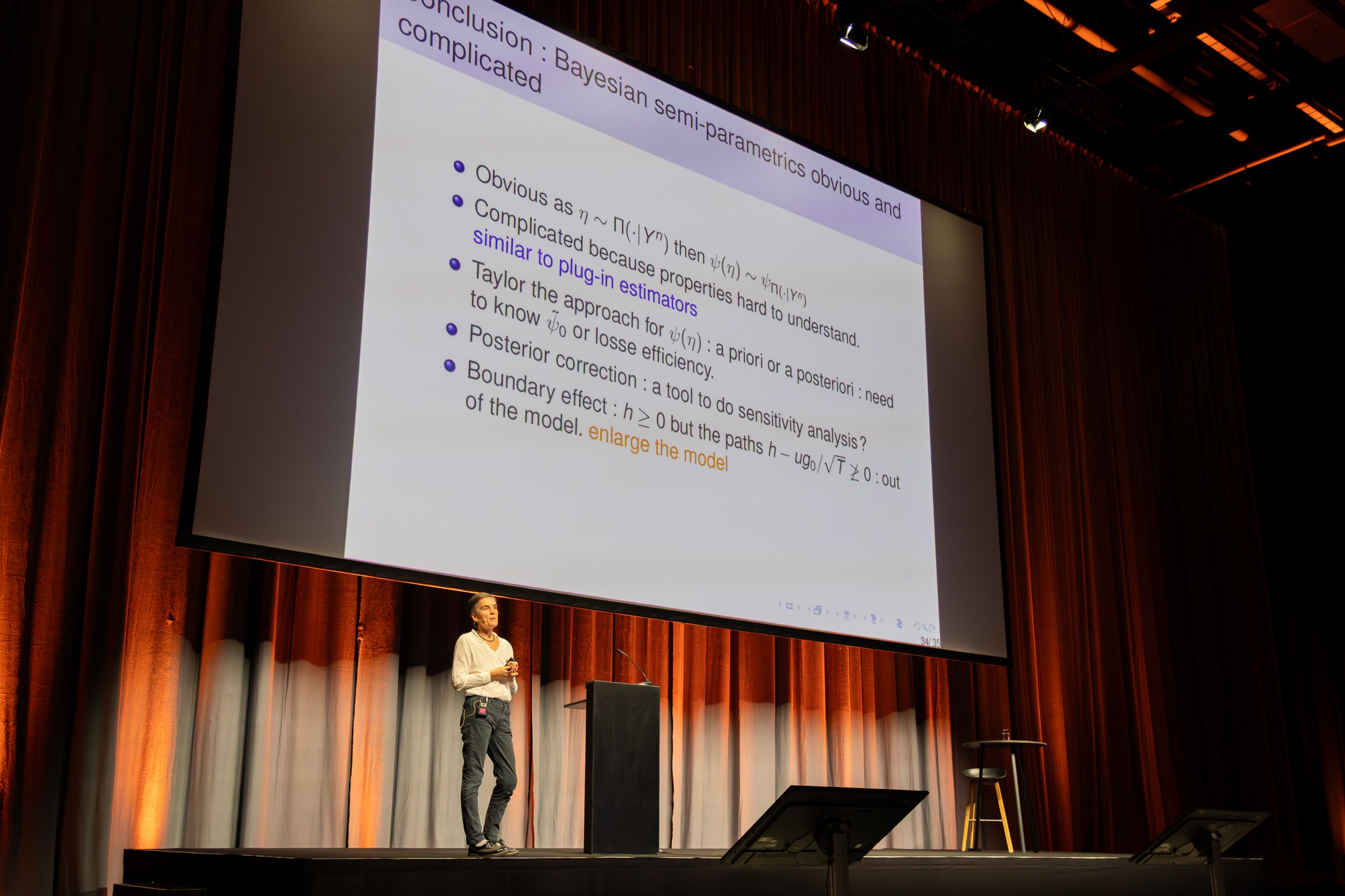 Judith Roussea standing on a stage in front of a large projected slide titled "Conclusion: Bayesian semi-parametrics obvious and complicated"