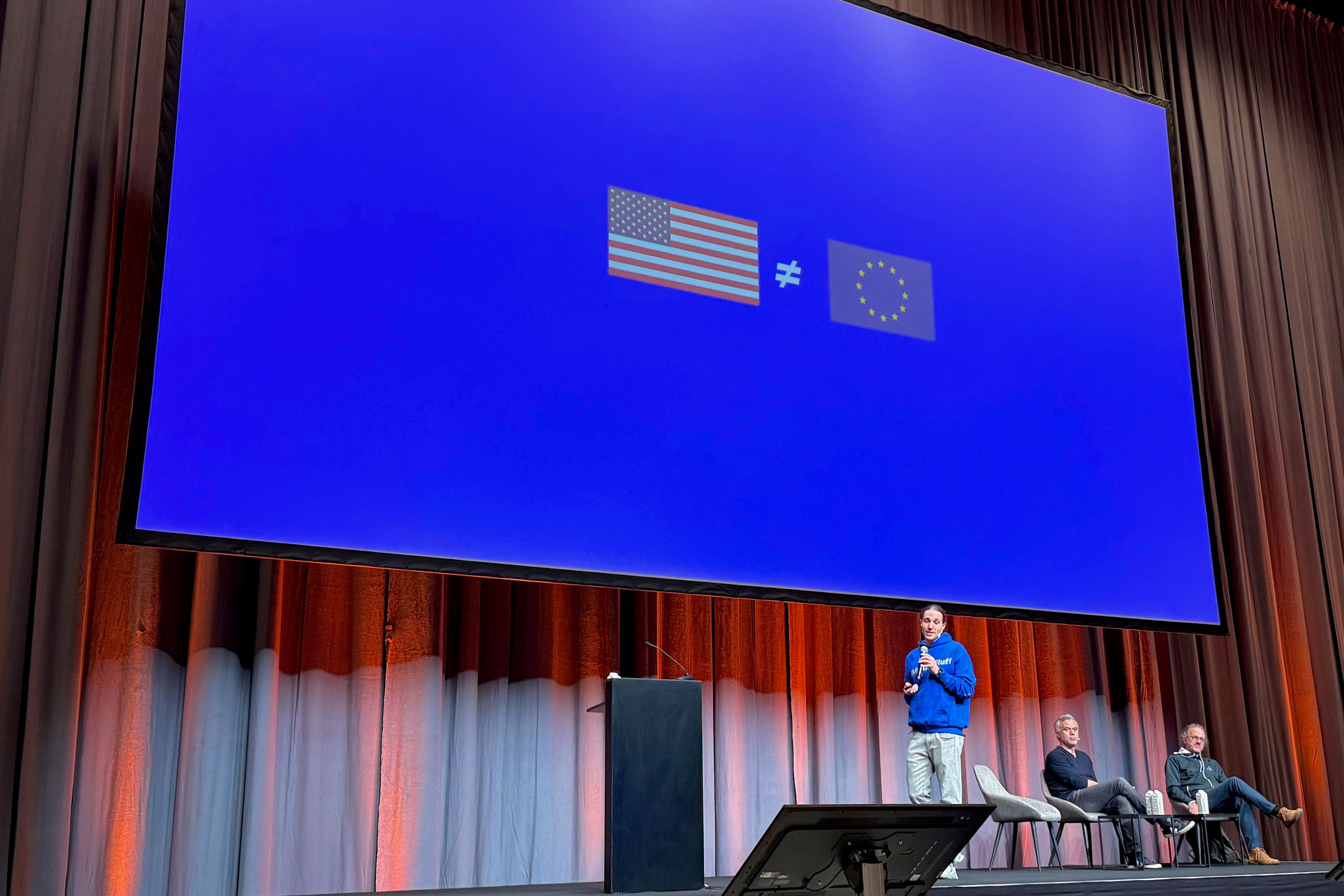 Johannes Otterbach on stage holding a microphone. On the screen behind him there is a US flag and a European flag, with the "not equals" sign in between them. Two people sit behind Otterbach on the stage. 