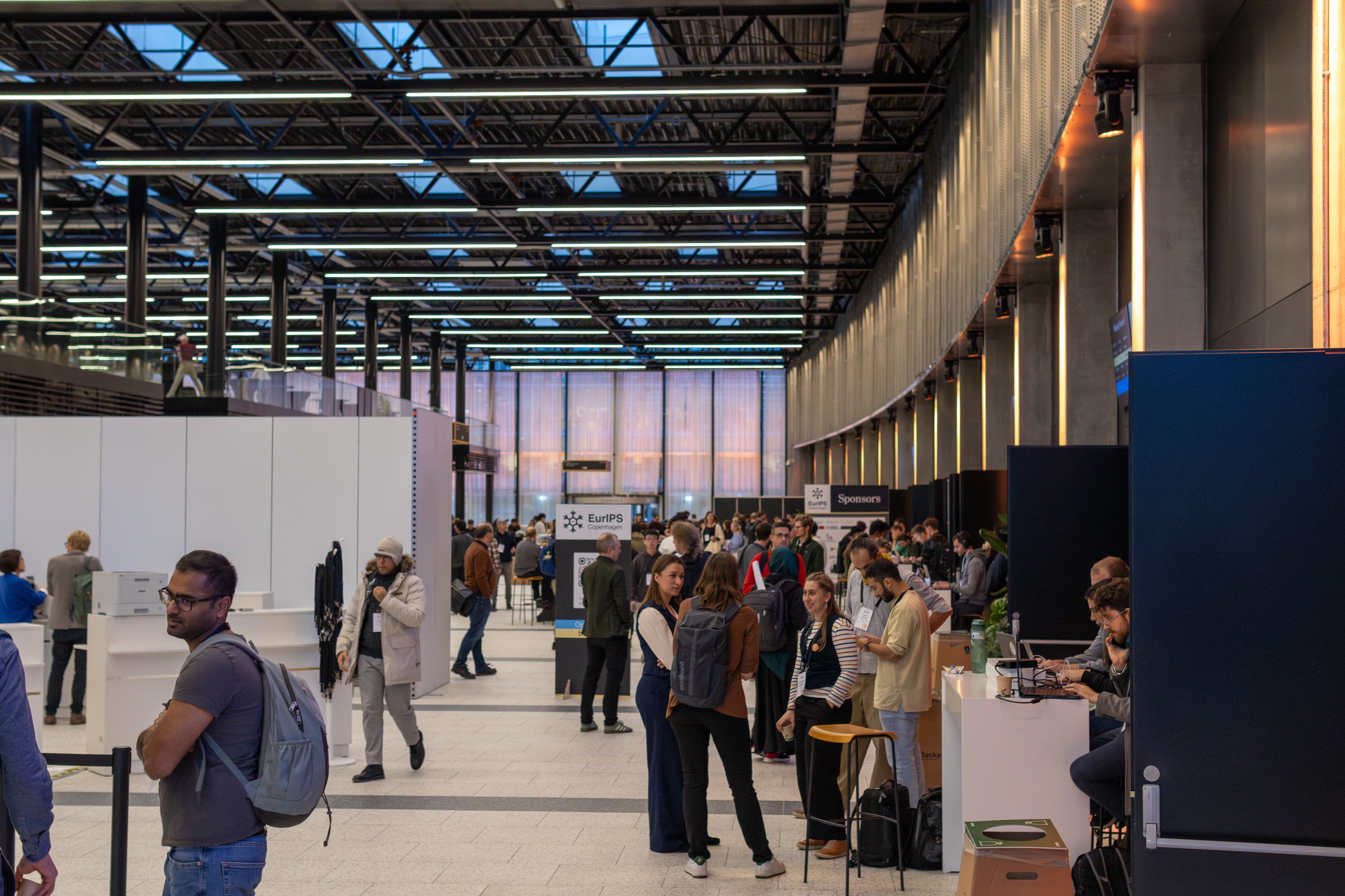 A large hall with high ceilings. People are dotted around in groups, talking to one another. A sign in the background reads "EurIPS".