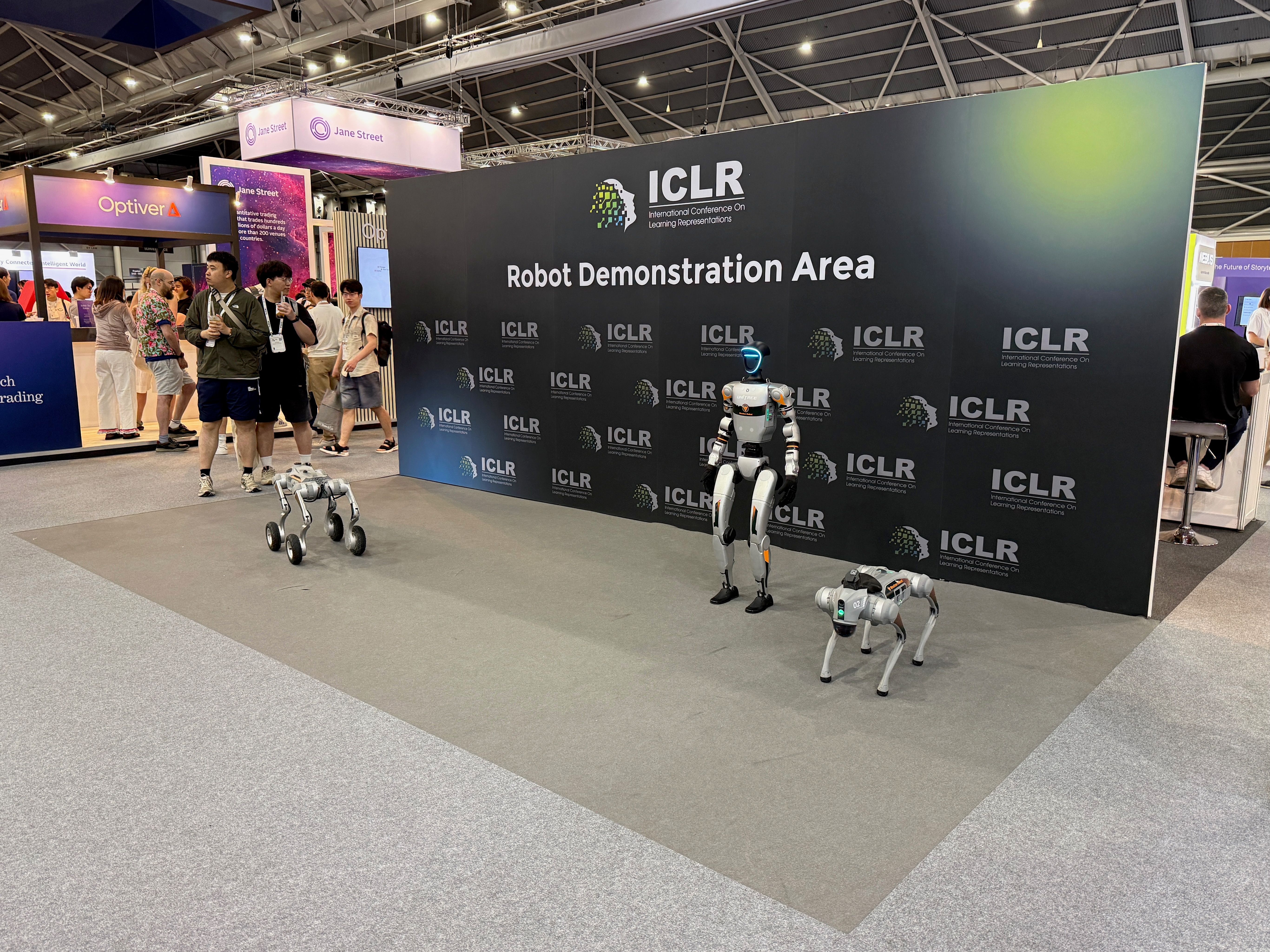 Three robots in front of a sign that reads 'Robot Demonstration Area'. One has four wheels on legs, one is a humanoid, and the third is a dog-style four-legged robot. 