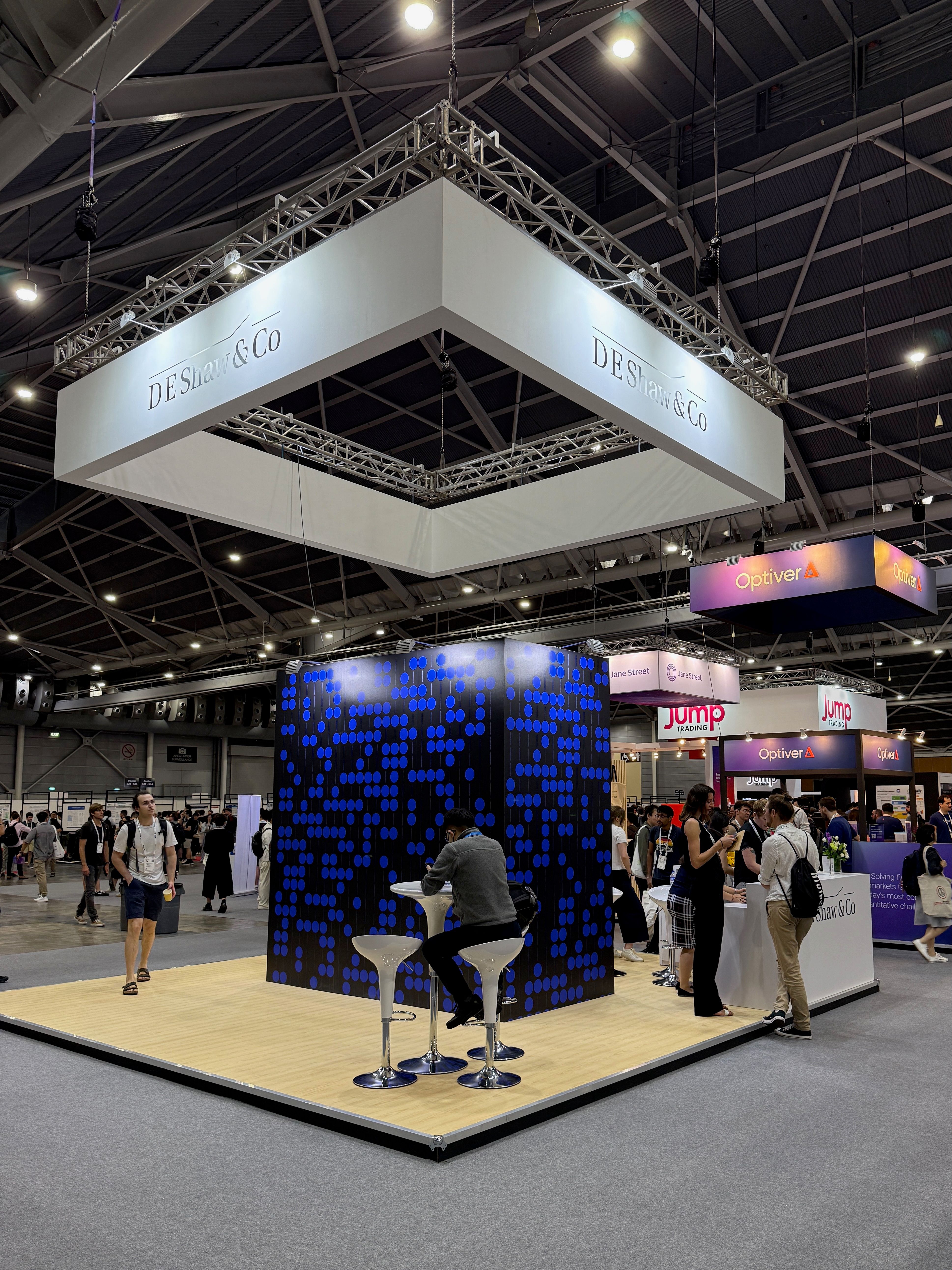 A large hall with sponsor booths. Visible are booths for D.E Shaw & Co, Optiver, Jane Street, and Jump Trading
