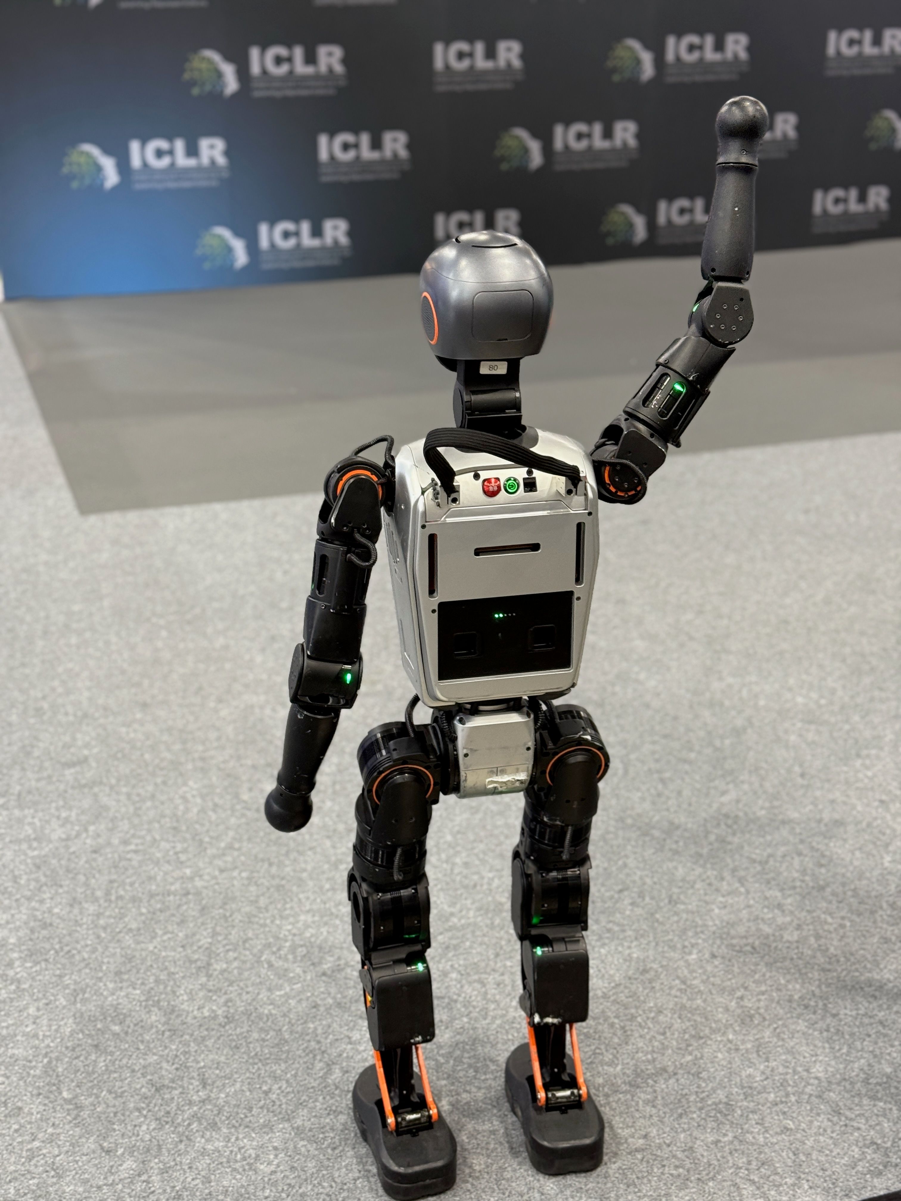 A humanoid robot 'faces away' from the camera, towards a wall tiled the ICLR logo, and 'waves'