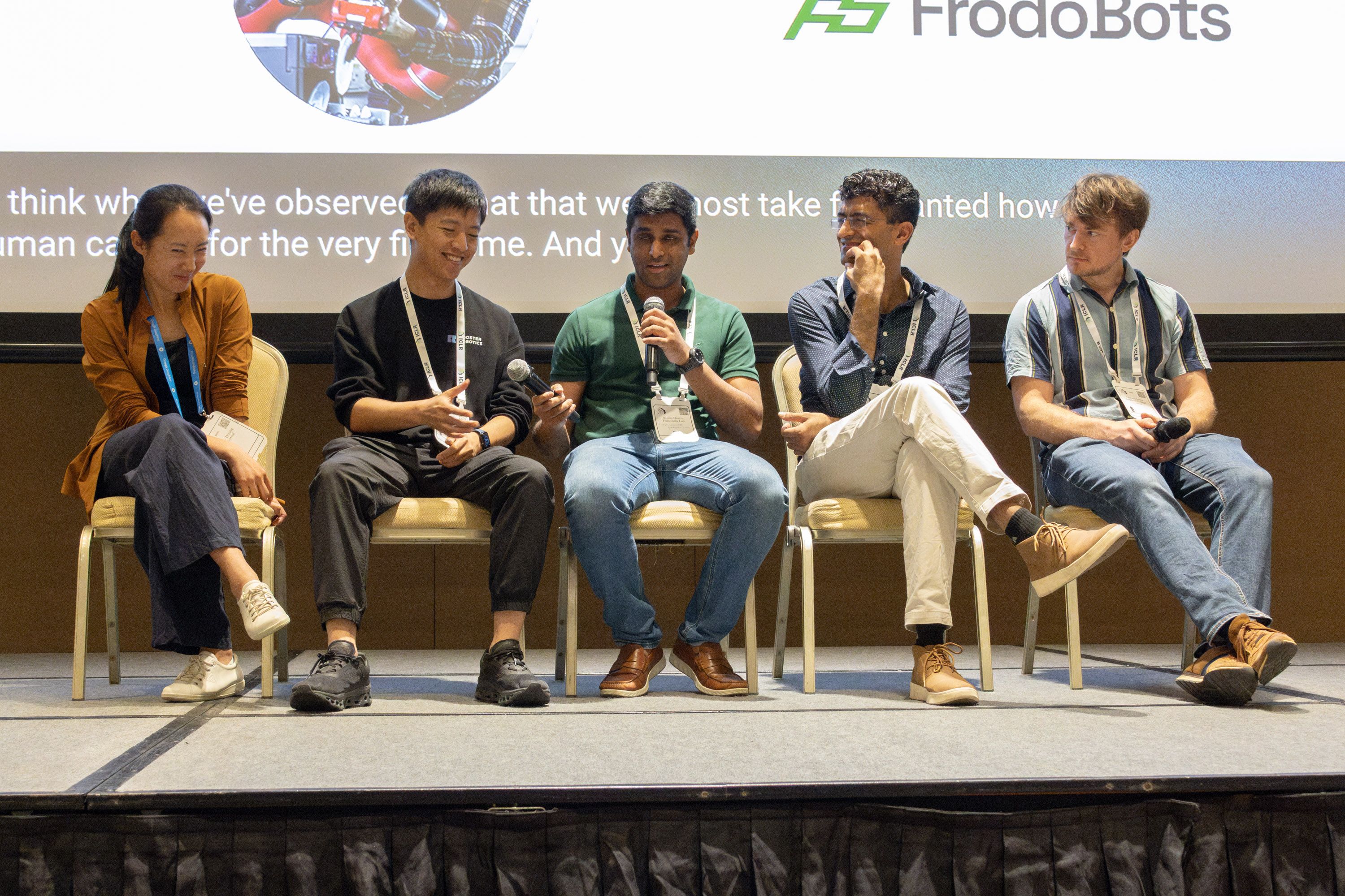 Sandy Huang, Chaoyi Li, Niresh Dravin, Animesh Garg, and Edward Johns sit on chairs on a stage, facing the audience. Dravin is speaking into a microphone while passing another microphone to Li. Most of the panelists are smiling. 