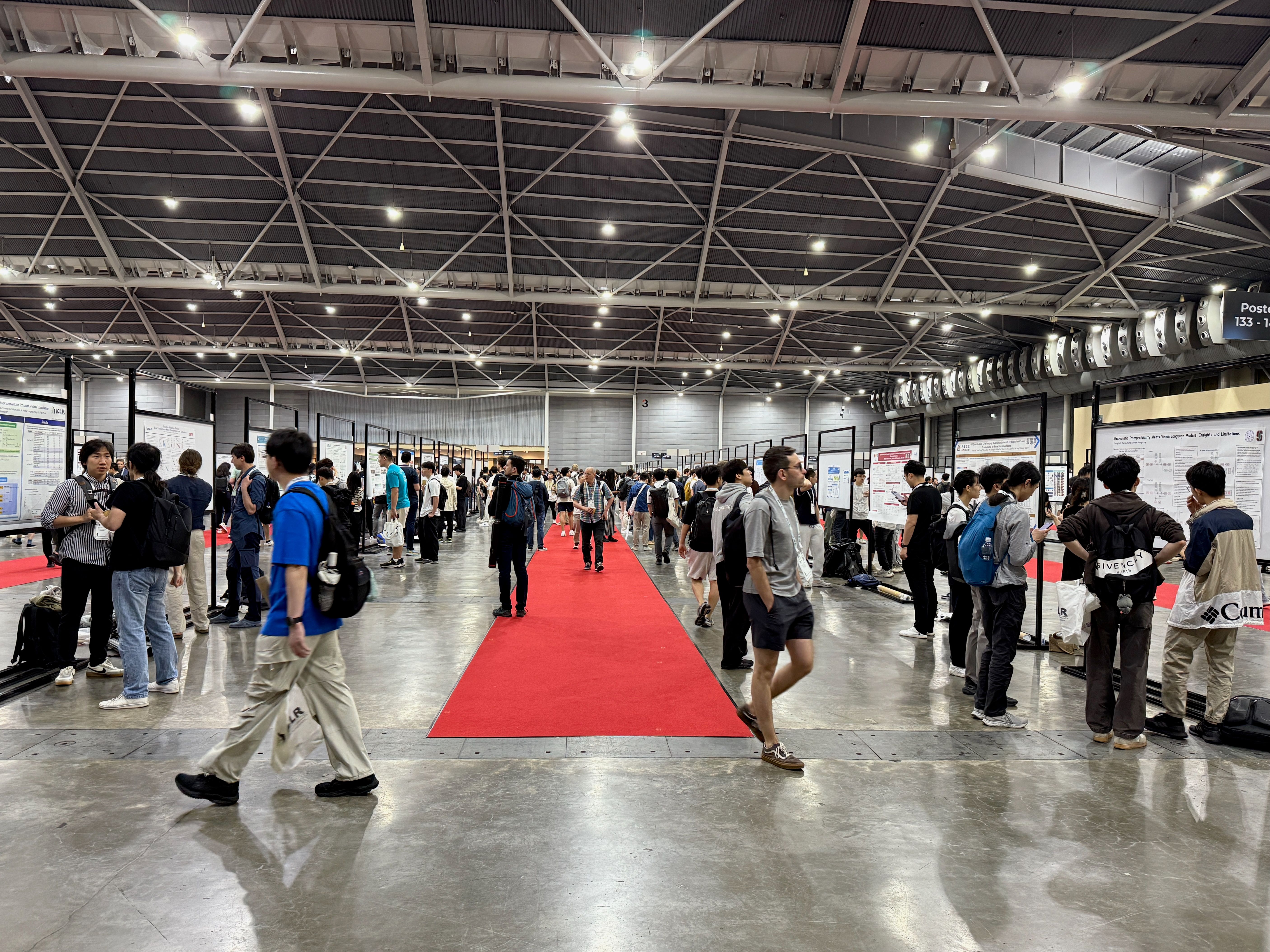 A large hall, with lots of people walking around. A red carpet runs through the middle of lines of academic paper posters.