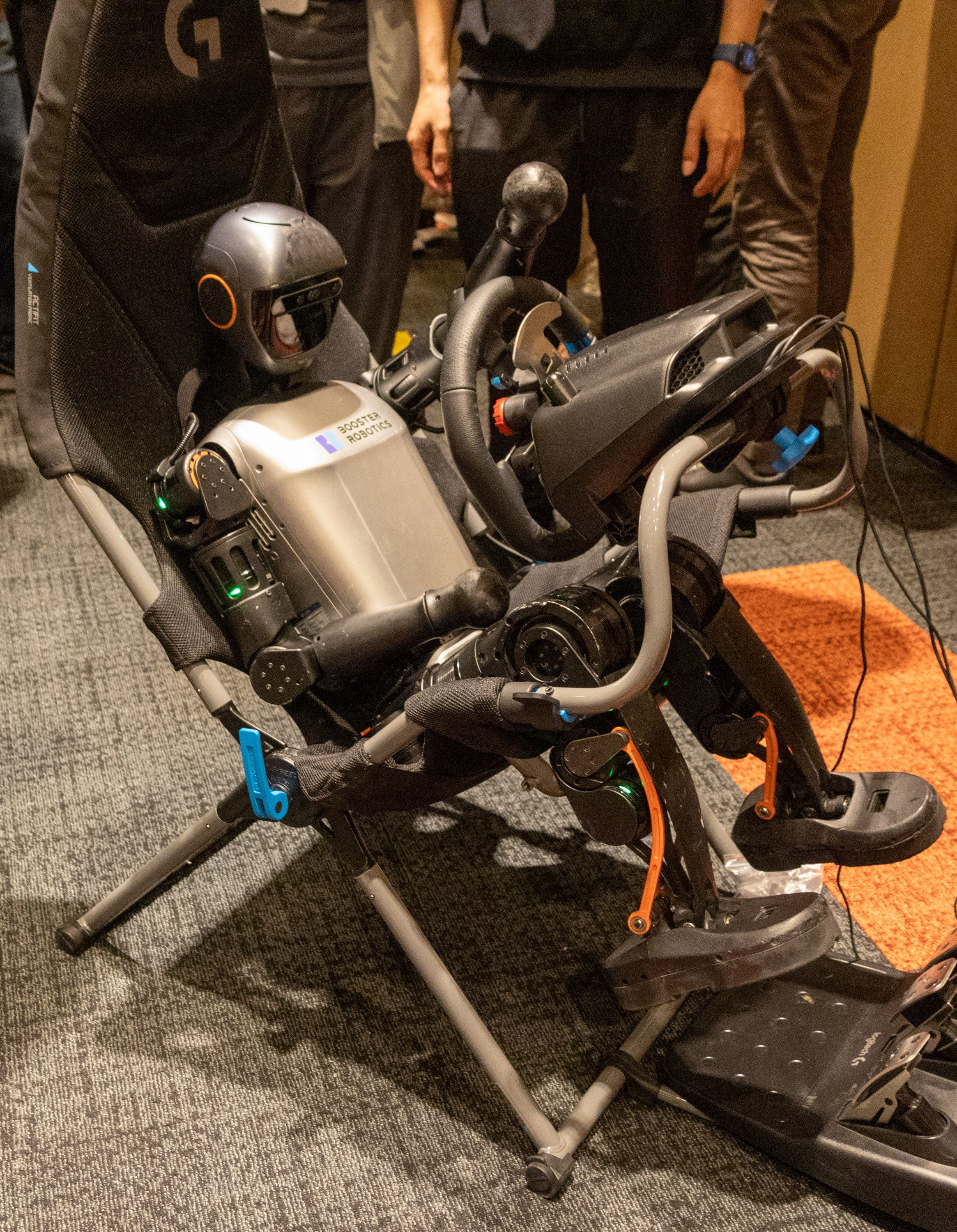 A child-size humanoid robot sits in a reclining chair behind a steering wheel and pedals. The robot does not have hands with fingers and cannot actually manipulate the steering wheel, but the photo is staged to make it look as if it can. 
