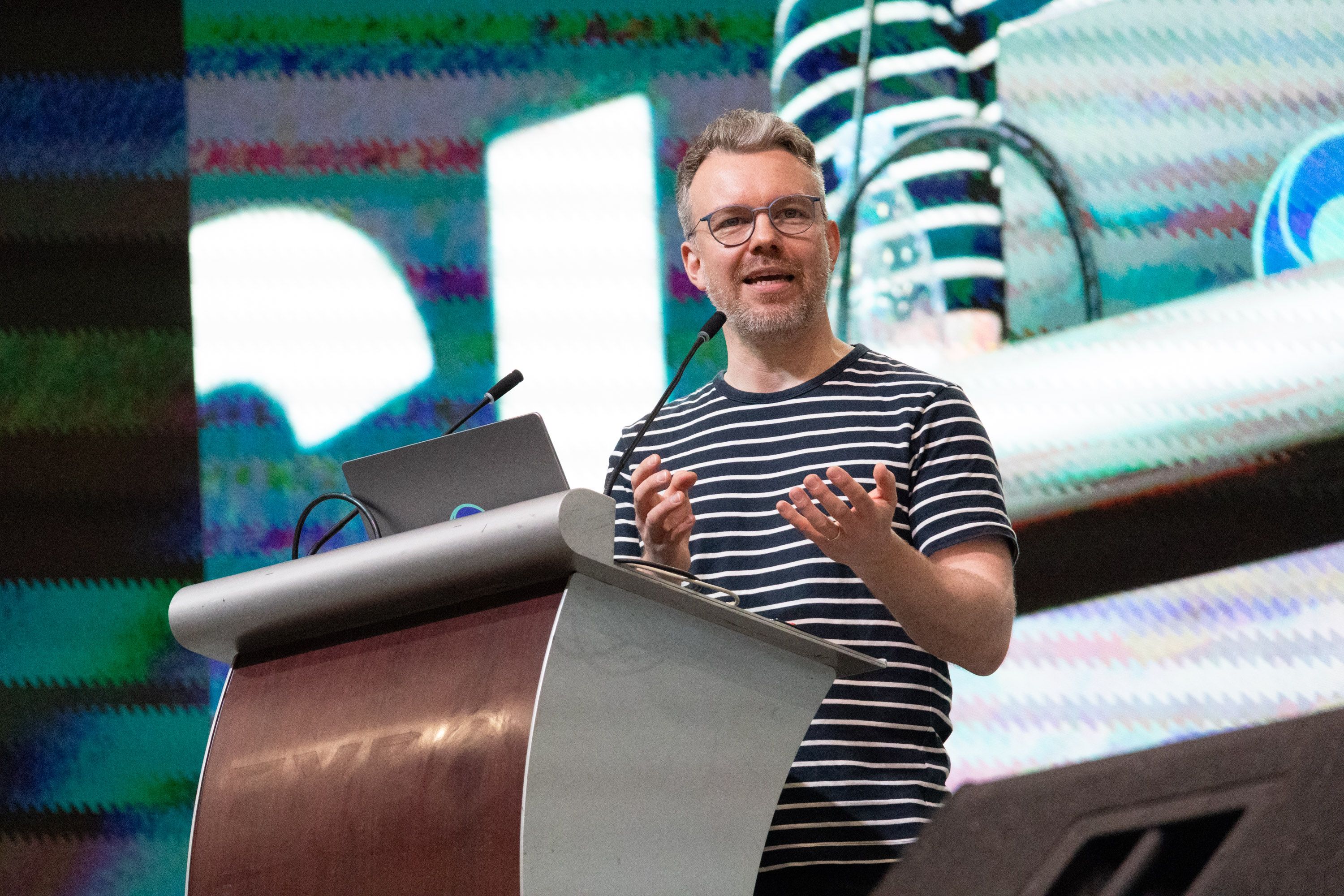 Tim Rocktäschel standing behind a lectern, addressing the audience, holding both hands in front of him in an expressive gesture. 