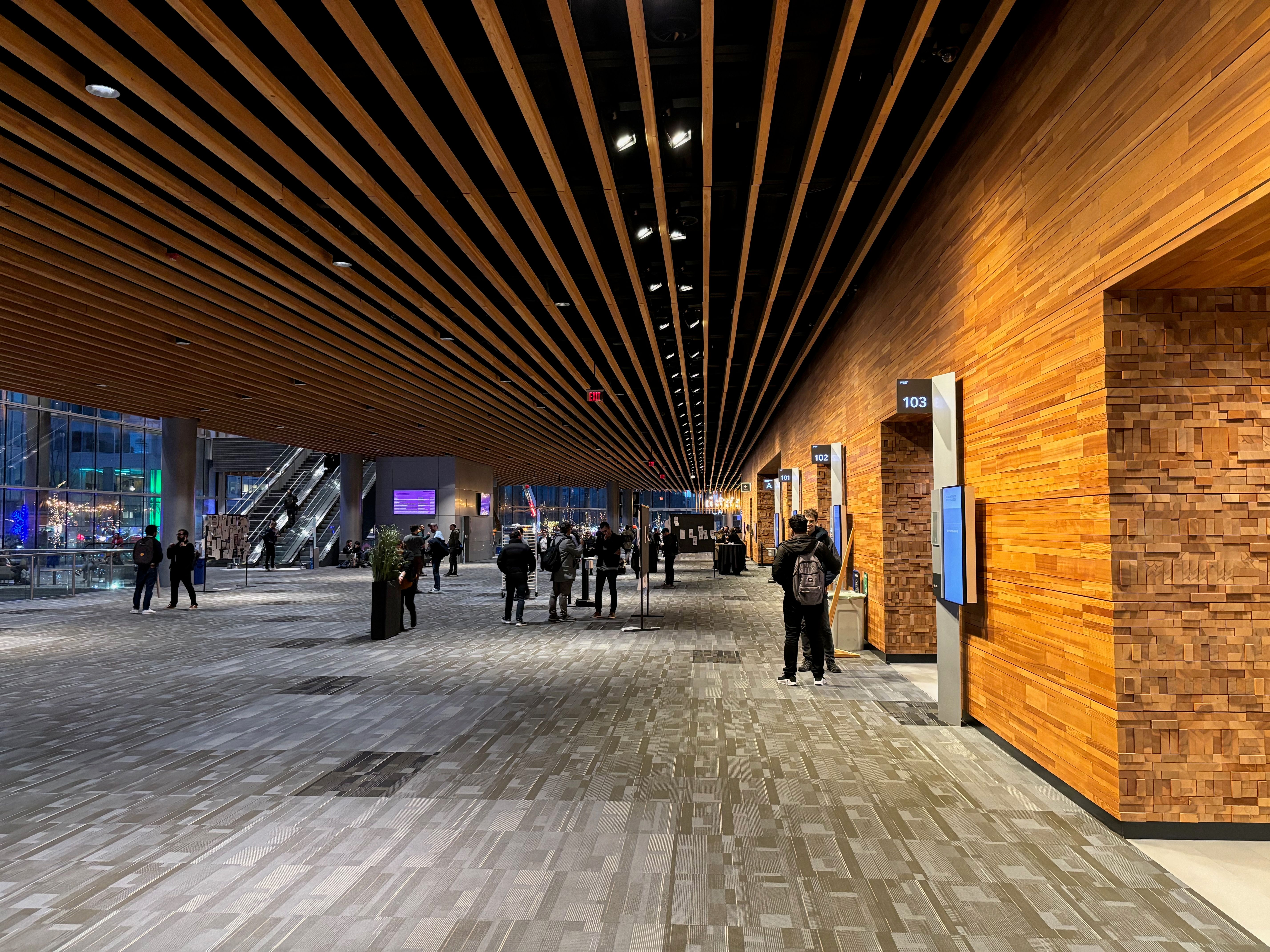 "The interior of a conference centre. A few researchers are walking around and talking to each other. Grey carpets, wooden slats on the ceiling. "